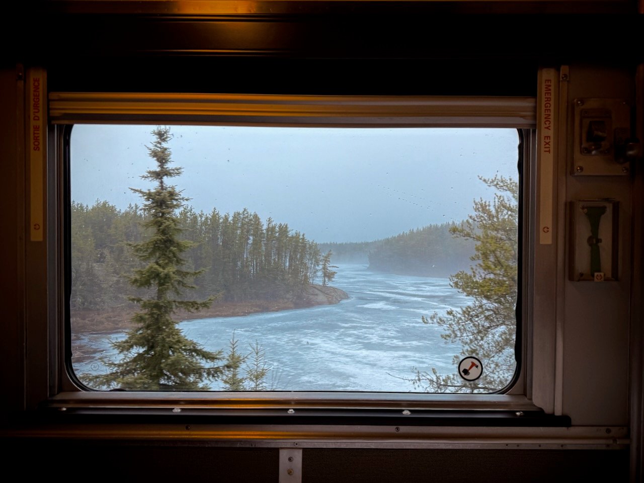 View from a train window of a frozen river and pine trees in a snowy landscape.
