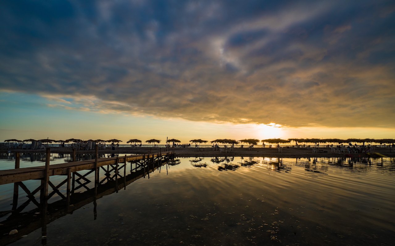 Sunset over a tranquil beach with a wooden pier and umbrellas reflecting on the water.
