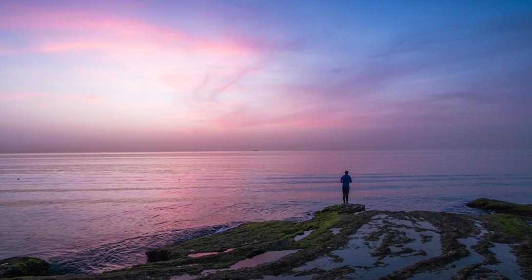 A person standing on a rocky shore at sunset, gazing at the calm sea under a pink and blue sky.