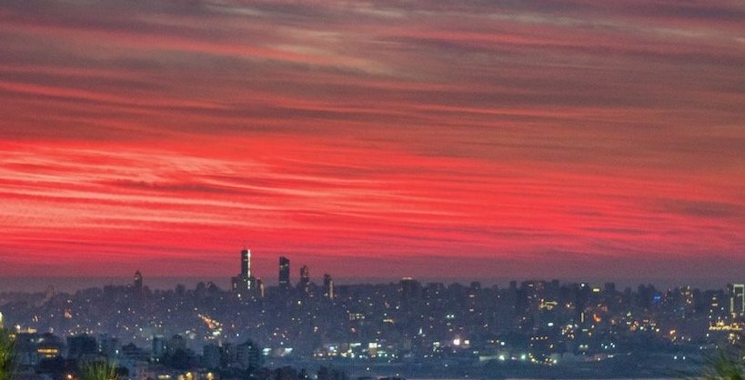 A cityscape at dusk with a fiery red sky over the skyline and illuminated buildings.