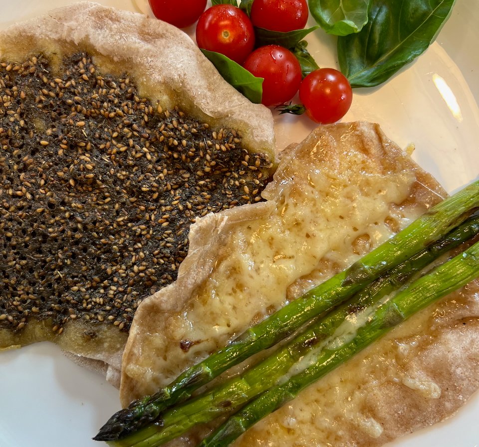 Fresh manakish with za'atar, cheese, grilled asparagus, cherry tomatoes and basil leaves on a white plate