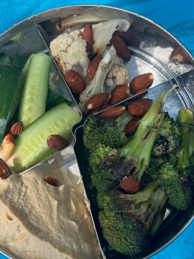 Snack box with hummus, fresh cucumbers, broccoli, cauliflower, and almonds on blue cloth
