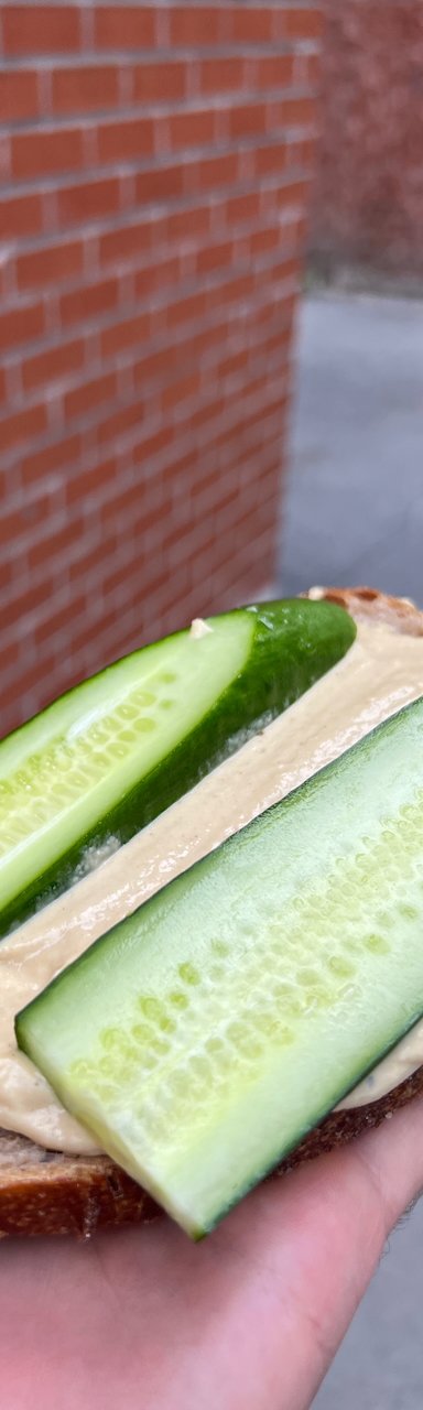 Hand holding slice of bread topped with hummus and fresh cucumber outdoors
