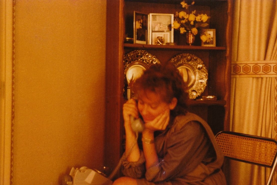 Woman on rotary phone in vintage room with ornate plates and flowers.
