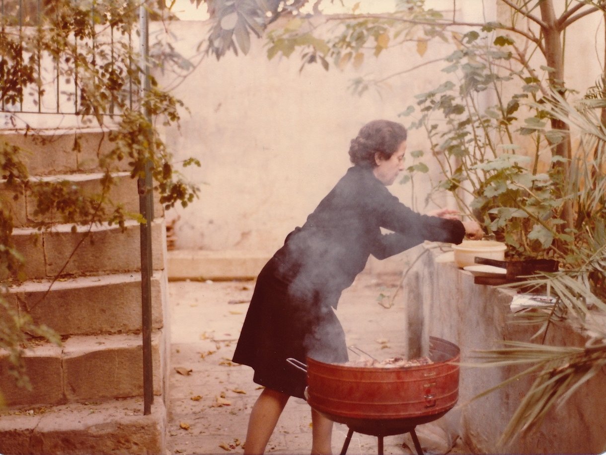 A woman cooking outdoors by a barbecue in a garden, surrounded by greenery and plants.
