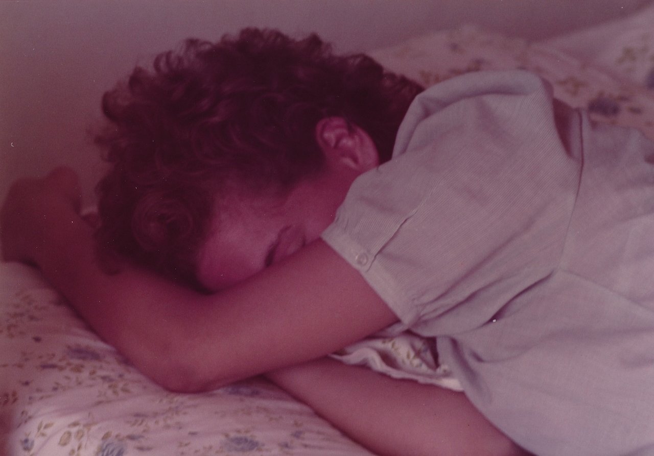 Person sleeping on floral bed with arms crossed, wearing a light shirt.