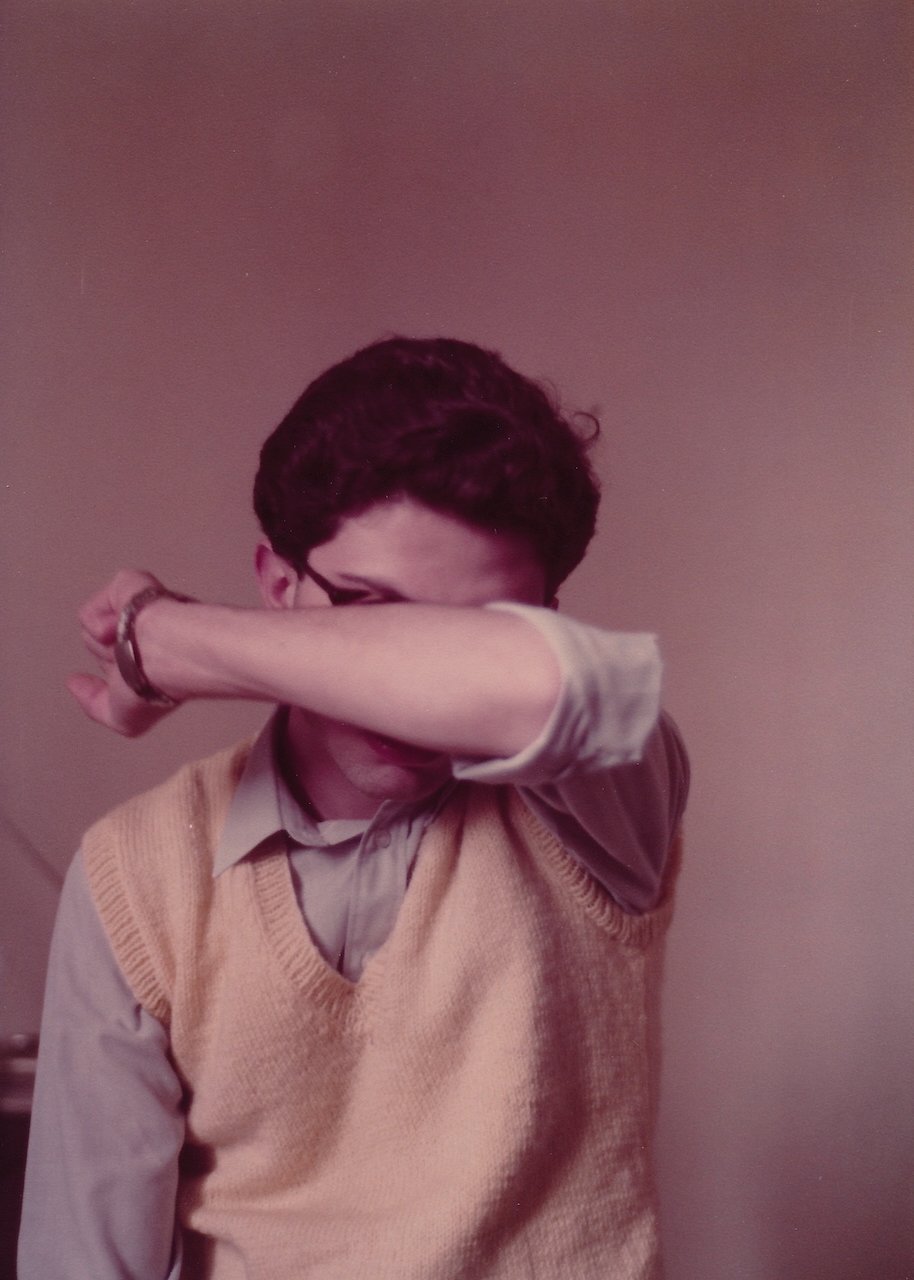 Person with glasses covering face, wearing a yellow sweater vest.