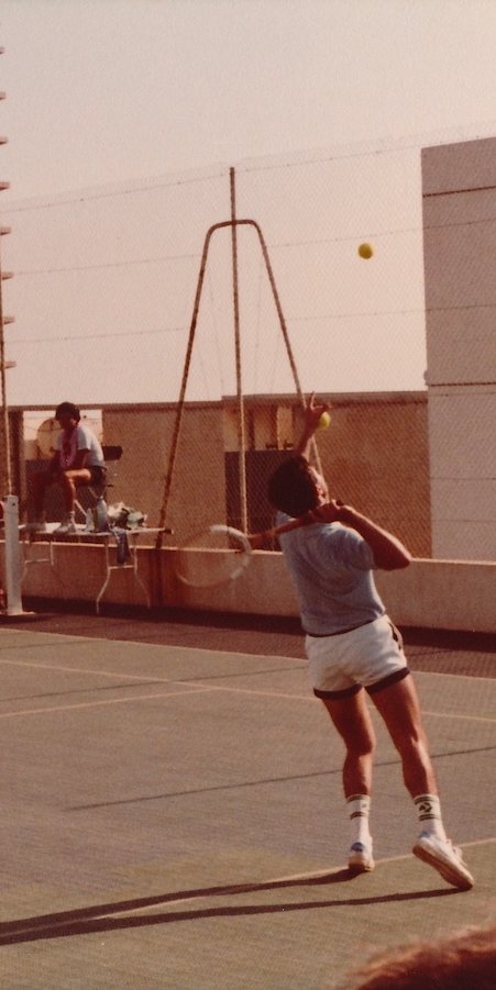 A player serves a tennis ball on an outdoor court, with a spectator seated in the background.