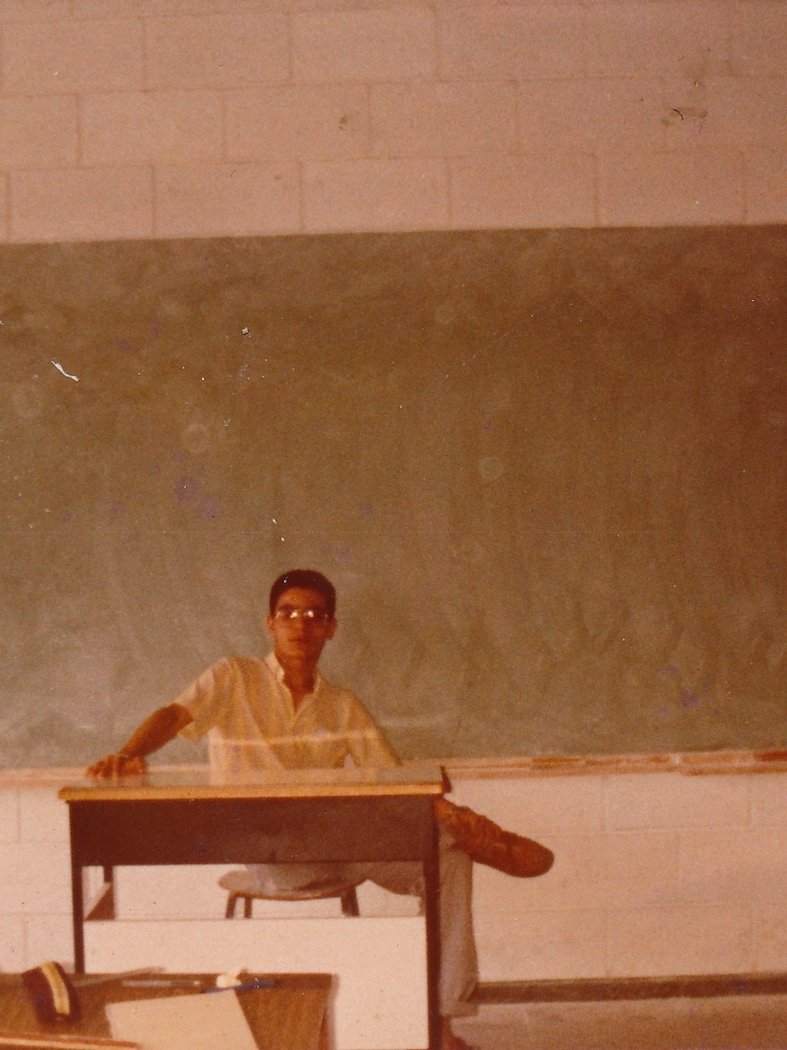A young man sitting at a desk in a classroom, wearing glasses and a white shirt, with a chalkboard in the background.
