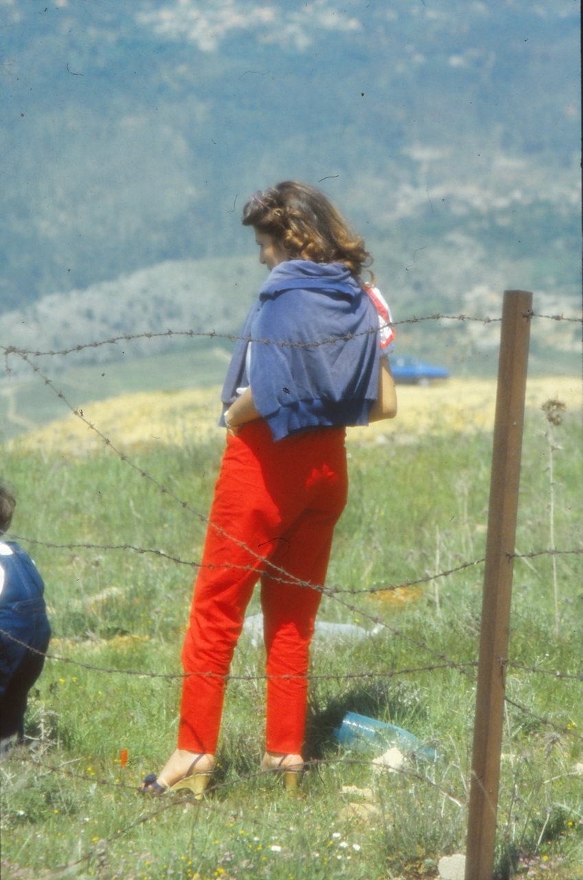 A woman in red pants and a blue sweater stands in a grassy field, with barbed wire in the foreground and a blurred landscape in the background.