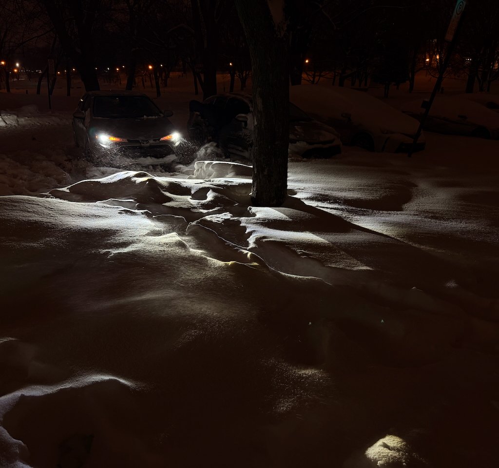 Car headlights illuminating snow-covered terrain and tree at night